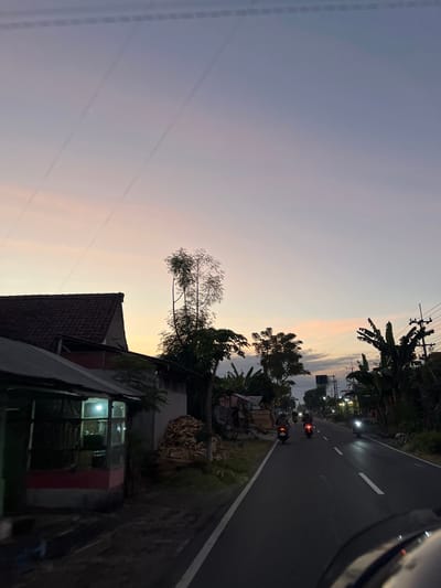 This is a vertical, eye-level shot taken from a moving vehicle on a road at dusk. The scene depicts a rural or semi-rural setting in Purworejo, Indonesia. The sky displays a gradient of soft purples and oranges, indicating sunset or twilight. Several power lines are visible crisscrossing the sky.
On the left side of the image, a tiled-roof building with illuminated windows suggests a dwelling or small shop. Adjacent to it, a leafless tree branches out towards the sky. Further down the road, there are banana trees and other vegetation, characteristic of tropical landscapes.
The road itself is asphalt with white lane markings, and it curves gently to the right. Several motorbikes are visible on the road, some with their tail lights illuminated, suggesting they are in motion. Streetlights are also present along the side of the road, casting a warm glow. There is a small roadside stall with some piled-up materials, possibly wood or agricultural produce. The overall mood is peaceful and serene, capturing the end of the day in a tranquil Indonesian landscape. No visible text is present in the image.