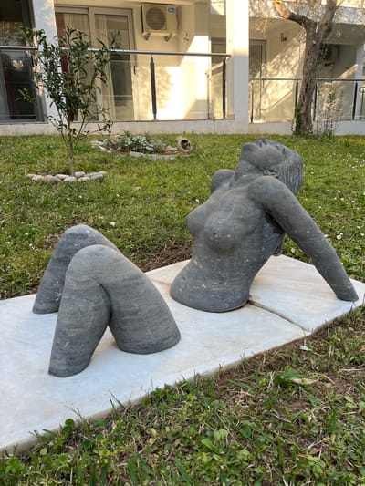 A partly-buried, grey, stone sculpture of a woman lies on white marble or stone slabs set into the grass. The sculpture is split into two pieces, a torso with one arm raised, and two legs bent at the knees. The location appears to be outdoors, near a building with large glass windows and doors. The ground is covered in grass and small white flowers. The image was taken in Brežine, Montenegro.