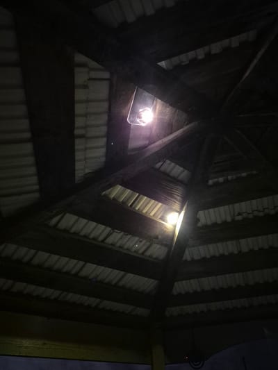 This is a low-angle, close-up, indoor shot of the roof structure of a building, likely a pavilion or gazebo, in Sars, Russia. The roof is constructed with dark wooden beams radiating from a central point and supporting corrugated metal roofing panels. Two bright light fixtures are visible, one near the top center and another lower and to the left, casting strong light and lens flare. The overall lighting is dim, with shadows obscuring much of the detail within the structure. The image does not contain any people or depict any actions or interactions.