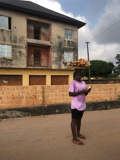 The image depicts a street scene in Ihiala, Nigeria, featuring an adult female street vendor in the foreground. The vendor, positioned centrally, is a dark-complexioned woman wearing a light purple short-sleeved t-shirt, black shorts, and red flip-flops. Visible sweat stains appear on her t-shirt. She carries a flat, circular tray on her head, laden with numerous small, yellow, plastic-wrapped items, likely food products for sale. In her hands, she holds a small, light-colored object, possibly a smartphone or a small container. She stands on an unpaved, light brown dirt or gravel road.
Behind the vendor is a low, unpainted concrete block wall of light orange/tan color. Beyond this wall, to the left, stands a multi-story building with a dark, sloping roof. The building's upper facade is weathered, with light beige plaster showing significant dark gray streaking and discoloration. The ground floor of this building features a yellow-painted section with three closed brown roll-down shutters. The upper floors have white-framed windows with reflective glass. Two central openings on the upper floors are covered by heavily rusted, reddish-brown metal security bars, with a wooden door visible behind the lower set of bars. To the right of the building, a dark utility pole with wires is visible against a partially cloudy sky. Green foliage from trees is present further in the background on the right.
