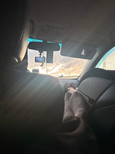 The image, oriented vertically, captures the interior of a car looking towards the front windshield. Strong, golden sunlight streams through a cloudy sky, creating a bright glare and lens flare across the upper and central parts of the frame.
On the right side, a person's forearm and hand are visible, wearing a light-colored, possibly knitted or ribbed, glove or sleeve. The hand appears to be resting or interacting with something near the front passenger area, though the specific activity is unclear.
The car's interior features include the headliner, a driver's side sun visor with a small mirror, and a rearview mirror. Mounted near the rearview mirror are several devices: a dashcam, a smartphone or GPS device displaying information, and another small electronic accessory. An air freshener, clearly labeled "areon X", hangs from the rearview mirror. Parts of the dashboard and a black seat are also visible.
The bright, low-angle sunlight suggests the time of day is either early morning or late afternoon/early evening. The sky is heavily clouded but the sun is breaking through. The scene is set inside a vehicle, with the provided context indicating the location is Alexandrovskoye, Russia. No specific emotions are discernible.