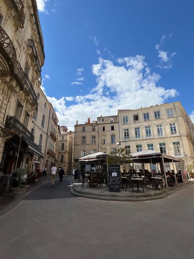 The image depicts a street scene in Montpellier, France, characterized by tall, weathered buildings with multiple windows and ornate wrought-iron balconies. The sky is bright blue with scattered white clouds. Along the street, outdoor seating areas of cafes or restaurants are visible, furnished with tables and chairs. Some people are walking on the pavement. The street is paved and shows signs of use. Chalkboard menus or signs are also present.