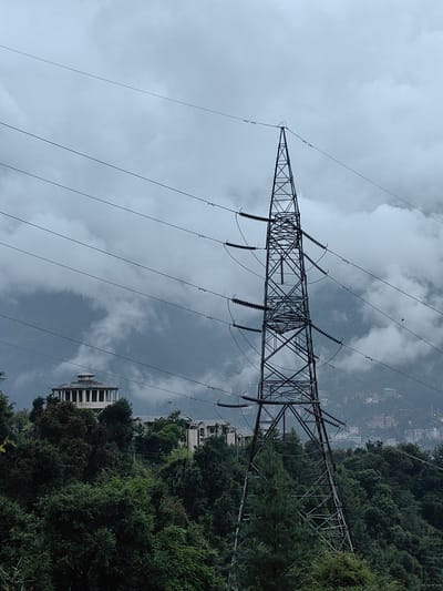 The image depicts a high-voltage electricity pylon situated on a hillside, overlooking a city and misty mountains. The pylon is a tall, lattice-steel structure with multiple crossarms supporting thick power lines. Several power lines stretch across the frame, disappearing into the cloudy sky. On the left side of the frame, a circular building with pillars and a domed roof is visible, nestled amongst dense green trees. Further in the background, parts of a city with buildings are visible, partially obscured by mist. The overall atmosphere is overcast, with the sky filled with heavy clouds, suggesting a damp or rainy day. The location is identified as Chhyal, India. There are no people or animals visible in the image.