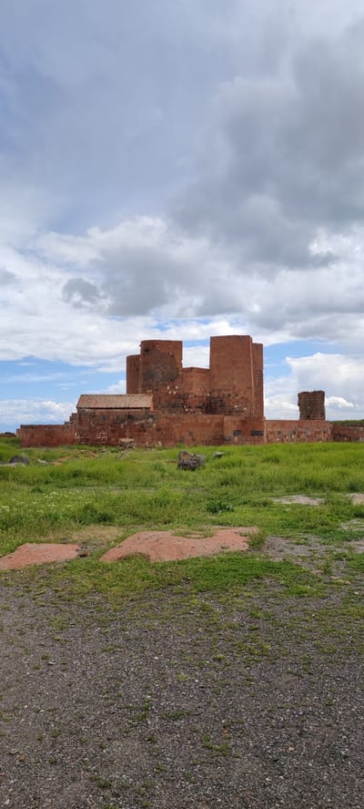 The image shows the Dashtadem Fortress in Armenia, situated against a backdrop of a partly cloudy sky. The fortress, constructed of reddish-brown stone, comprises multiple connected structures, some taller and more prominent than others. One section features a sloping roof. The structure is partially dilapidated.
The foreground is dominated by gravel and patches of green grass intermingled with red, rocky soil. The vegetation appears sparse and uneven.