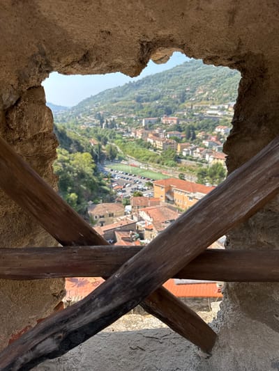 The image provides a view through a rustic, irregular opening in a stone wall, framed by weathered wooden beams. The opening looks out onto a sunlit, verdant hillside dotted with buildings. In the mid-ground, a collection of houses with terracotta roofs and cream-colored facades are visible, interspersed with trees. A paved area, appearing to be a parking lot, is situated below the houses, next to a green rectangular field, possibly a sports field. Further up the hill, more buildings are scattered amongst the trees, and the landscape extends to a hazy, distant mountain range. The overall scene suggests a view overlooking the town and surrounding valley of Dolceacqua, Italy. The stone structure in the foreground is rough and textured, with exposed mortar and irregular edges. The wooden beams are dark, aged, and appear to be structural elements. No people are visible in the image.