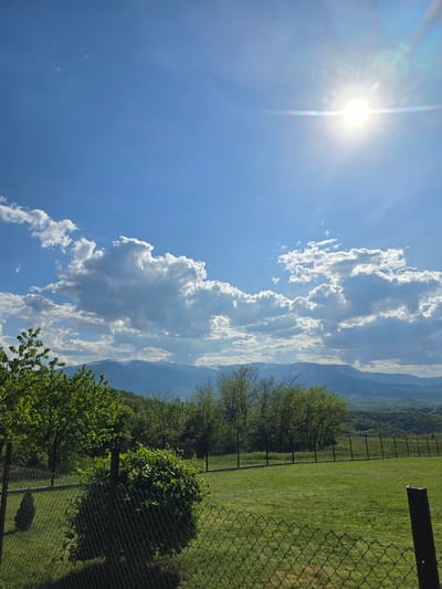The image shows a daytime landscape in Bokilovtsi, Bulgaria. In the top left, the sun shines brightly in a blue sky with scattered clouds. Vertically, there is a line of puffy white clouds extending towards the upper middle of the image, behind which indistinct mountains are visible. To the right, there is a green field with grass and some trees along a fence line. The fence is made of chain-link and there is a wooden post visible. The angle of the image is slightly rotated, giving a tilted perspective of the landscape.