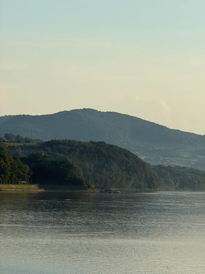 This is a landscape photograph of a body of water, likely a lake or river, with rolling hills covered in dense green trees in the background. The sky is clear and pale blue, with a slight gradient towards a lighter tone near the horizon. The water's surface is rippled, reflecting the light. On the shoreline, there are hints of human structures, including a road or path with some fencing, and what appear to be small buildings or structures nestled amongst the trees on the hills. The overall impression is a serene, natural scene. The location is in Lipie, Poland.