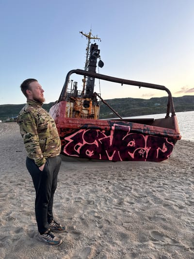 A man stands on a sandy beach in Teriberka, Russia, looking to his left. He is wearing a camouflage jacket, dark pants, and athletic shoes. Behind him is a rusty, dilapidated vessel covered in pink graffiti. The vessel appears to be beached on the shore. The sea is visible behind the vessel, and rolling hills with some sparse vegetation and buildings can be seen in the distance. The sky is clear and light blue, suggesting it is either early morning or late evening. The overall scene depicts a somewhat desolate and industrial seascape.