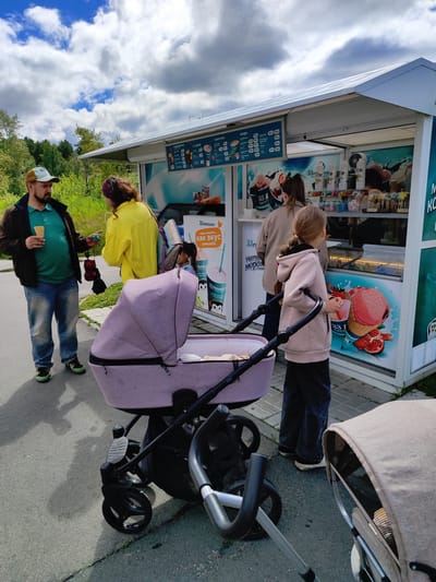 In the city of Snezhinsk, Russia, a scene unfolds outside an open-air ice cream kiosk under a cloudy sky. A man wearing a brown jacket, green shirt, and blue jeans stands to the left, holding an ice cream cone and a small, brown umbrella. Beside him, a woman in a bright yellow jacket faces away from the camera, holding a small, yellow object. In front of them, a light purple stroller with a black frame is positioned centrally. To the right of the stroller, a young girl with braided hair, wearing a pink hooded sweatshirt and dark pants, looks towards the kiosk. Behind her, another woman in a beige sweatshirt stands facing the kiosk. A second, tan stroller is partially visible on the far right. The kiosk features a menu board displaying prices and images of various ice cream products and drinks. The ground is paved with asphalt and some cobblestones near the kiosk. Greenery and trees are visible in the background.