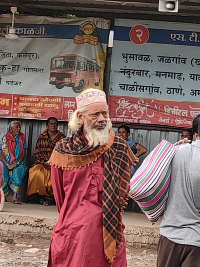 The image depicts an outdoor scene, likely a bus stop or market area, in Malkapur, India. In the foreground, an elderly man with a white beard and a white embroidered cap is the central figure. He wears a dark red kurta and a checkered scarf with orange and black patterns. To his left, three women are seated on a raised concrete platform. The woman on the far left wears a blue and pink sari with a red jacket. The middle woman is dressed in a dark brown and orange patterned sari. The woman on the right is wearing a blue scarf over a dark top. Behind them, a large banner with Marathi text and an image of a red bus is displayed. Text on the banner includes "ST" and lists several city names, suggesting a bus route or service information. Another banner below it features Marathi text and the English phrase "THE REAL RESULT OF PRINTING". In the background, more people are visible, and a person in a grey t-shirt and brown pants stands to the right, holding a striped bag with green, pink, white, and purple stripes. The environment appears somewhat weathered, with visible concrete structures and a corrugated metal wall in the background. The overall impression is one of a busy public gathering place.