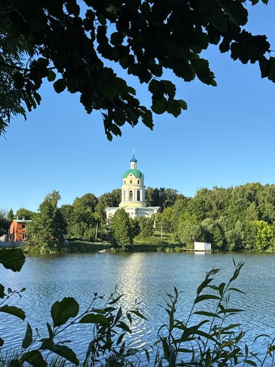 The image shows a serene view of the Church of the Assumption of the Blessed Virgin Mary in Grebnevo, Russia, situated on the bank of a tranquil lake. The church, with its distinct green dome and yellow bell tower, is nestled amidst lush green trees, creating a picturesque backdrop. The foreground features the calm, rippling surface of the lake, reflecting the clear blue sky. Silhouettes of leaves from overhanging trees frame the top of the image, adding a natural touch to the scene. A red building is visible to the left of the church, and a small white structure stands on stilts in the lake on the right side. The overall atmosphere is peaceful and idyllic, typical of a summer day in a rural setting.