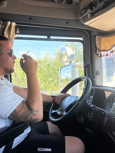 A man is driving a truck on a sunny day. He is wearing sunglasses, a white t-shirt, and black shorts. He has tattoos on his left arm and is holding a cigarette in his right hand. The truck's interior is visible, including the steering wheel, dashboard, and side mirror. Outside the truck, trees and a blue sky can be seen. The location is stated to be in the city of Lyra, Greece.