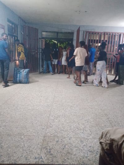 A group of people stands near a gate or doorway in what appears to be a public building. The light is dim and the picture grainy. People are scattered, some looking out the opening, others turned to the side. A striped bag rests on the floor near the left side of the image. The floor is made of light-colored square tiles. The scene is casual and potentially a gathering of individuals.