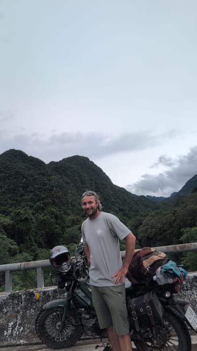 A man with light brown, wavy hair and a beard, wearing sunglasses on his head, a grey t-shirt with a white compass graphic, and olive green shorts, stands next to a dark green motorcycle. The motorcycle is loaded with luggage, including a large backpack and two saddlebags. A helmet rests on the handlebars of the motorcycle. The man has his right hand on his hip and is looking at the camera with a smile. The motorcycle is parked on a bridge with a concrete barrier and railing. In the background, lush green mountains covered in trees rise up against a cloudy sky. The location is Thượng Trạch Commune, Vietnam.