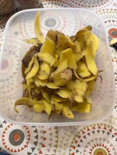 A clear plastic container filled with potato peels is placed on a table. The container is positioned centrally in the frame, and the peels are piled high within it. The potato peels are mostly yellow with some brown skin still attached, indicating they have been recently peeled. Some of the peels are curled, while others are laid flat. The table beneath the container has a patterned tablecloth featuring circles and triangles in various colors, including red, orange, grey, and green. The background is slightly out of focus, but a blurry object wrapped in plastic, possibly more potatoes, can be seen in the upper left corner. A portion of a knife with a blue handle is visible on the right side of the frame, resting on the tablecloth. The lighting appears to be natural, casting subtle shadows on the peels. The image does not provide any specific indicators of a location in the city of Морозовка, Russia.