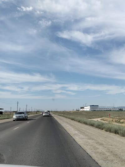 The image depicts a view from a vehicle traveling on a paved road in a semi-arid landscape. The sky is a vibrant blue with wispy white clouds, suggesting a clear and sunny day. The road ahead is straight and appears to be a highway with multiple lanes. Several cars are visible on the road, including a silver sedan in the foreground and white cars further ahead. The roadside is characterized by dry grass, sparse vegetation, and dirt. In the distance, a large industrial building with a white facade and blue accents is under construction, with cranes visible near it. Utility poles with wires line the side of the road, indicating infrastructure in the area. The overall setting suggests an industrial or developing region. The time of day appears to be daytime, likely afternoon, given the bright sunlight and clear skies. The location context is Ömirzaq, Kazakhstan. There is no text visible in the image.
