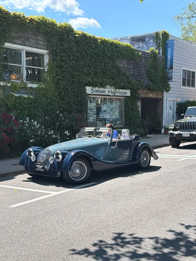 A dark blue convertible classic car is parked on a street in the Village of Sag Harbor, USA. The car has wire-spoke wheels and a prominent grille. A person wearing a blue shirt is seated in the driver's seat. Behind the car, a building is covered in dense green ivy, with a sign that reads "Simon Harrison" above a shop window. To the right of the building, another sign hangs vertically and reads "DEAR EVAN HANSEN". Further to the right, the rear of a dark green Jeep Wrangler is visible, with a license plate that reads "KLH-6420". The scene is set on a sunny day with a bright blue sky and scattered clouds.