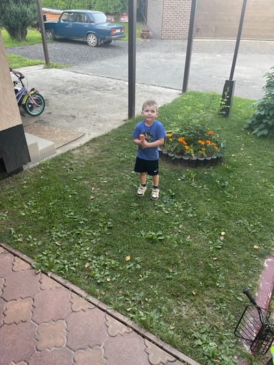 In this outdoor medium shot, a young boy with blonde hair is standing in a grassy yard, holding a small orange toy. He is wearing a blue t-shirt and black shorts, with white and black sneakers. The boy is looking towards the camera with a neutral expression. Behind him, to the left, is a flower bed with bright orange flowers and a dark grey border. Further in the background, a blue sedan is parked on a gravel driveway.
The scene appears to be a residential area. The grass is green and slightly uneven, with some scattered leaves and weeds. The ground is a mix of grass, gravel, and paved surfaces. A section of a brick building is visible on the far left, suggesting a house or other structure. A black metal bicycle basket is also visible in the upper right corner of the image. The lighting suggests it is daytime, likely overcast given the soft shadows. The overall impression is a casual, everyday moment captured outdoors. The location is identified as Савельево, Russia. There is no visible text in the image.