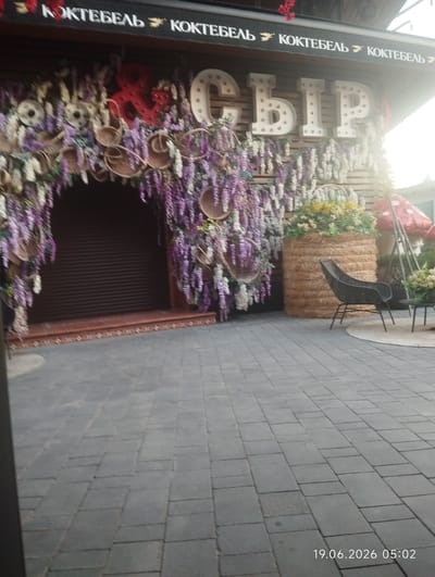 In an outdoor setting, the facade of a building features a large sign with illuminated white letters spelling "СЫР", which translates to "cheese" in English. The sign is decorated with what appears to be floral garlands, a red ampersand, and baskets. To the right of the sign, a large cylindrical hay bale is adorned with yellow and green flowers, next to which is a black woven chair and a small table. Behind these, a red mushroom-shaped decoration and a ladder are visible, as well as more floral arrangements. The ground in front of the building is paved with grey rectangular stones. A date and time stamp, "19.06.2026 05:02", is present in the lower right corner of the image. The location is identified as Kiparisnoe, Ukraine.