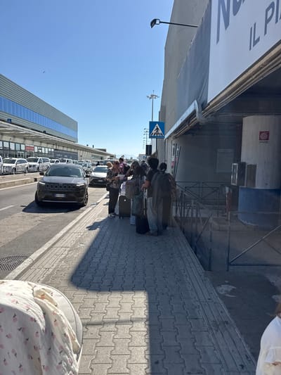 The image shows a group of people standing with luggage on a sidewalk in Cinisi, Italy. The sidewalk is paved with interlocking gray blocks. The people appear to be waiting.

A row of parked cars lines the street, and a black Jeep is closest to the viewer. The license plate is visible and reads "CY 189CB".

A modern building with a glass facade is on the left side of the street, with multiple visible storefronts. Above the building, the clear blue sky is visible. A pedestrian crossing sign is present. Above the sidewalk are industrial elements. The image is taken at an angle.
