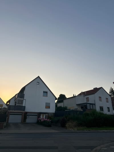 In Schöningen, Germany, an outdoor, low-angle, full shot shows two houses from a street at dusk. The sky is clear, with the sun setting on the left side of the frame. The houses are multi-story structures with white facades and dark roofs. The house on the left has a balcony with a dark railing and two garage doors on the ground floor. Pink roses are visible in front of the house. The house on the right has a red tiled roof and several windows with closed shutters. A rooster weather vane is perched on the roof of this house. The area in front of the houses is landscaped with bushes and trees. A street with gray asphalt is in the foreground.