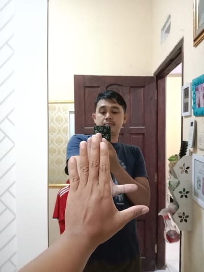 The image depicts an indoor mirror selfie taken in Brangsong, Indonesia. A male individual, appearing in his 20s or 30s, with short dark hair and a mustache, is reflected in a mirror. He is wearing a dark blue short-sleeved t-shirt and dark pants. He holds a black smartphone, featuring a white patterned case, in both hands at chest level, focusing his gaze on the device. The right hand of the photographer, belonging to the same individual, is extended into the immediate foreground, palm facing the camera with fingers adjoined.

The reflected background behind the man includes a partially open dark brown paneled wooden door. To the left of the reflected figure, a section of patterned beige wallpaper and a red garment with white stripes are visible. To the right of the reflection, a light-colored wall displays a series of mounted objects: an upper gold-framed picture, a light blue-framed picture, several white electronic or utility boxes, and a white decorative shelf with flower cutouts. This shelf contains a black comb, green containers, and a clear spray bottle with red liquid. A white-framed picture is positioned below the shelf. The visible walls are predominantly light beige or cream-colored.