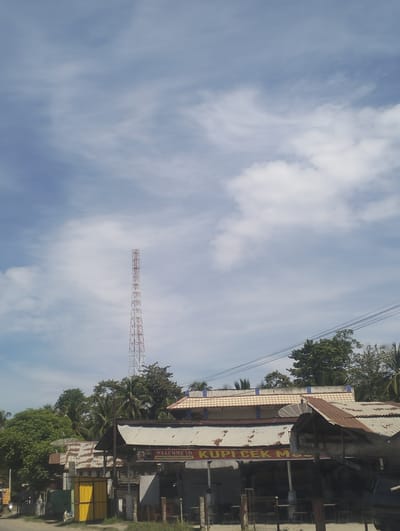 This is an outdoor, eye-level, vertical shot of a street scene in Grong-Grong, Indonesia. The sky is partially cloudy and appears to be a bright, sunny day, likely mid-morning or early afternoon. In the background, a tall, red and white lattice telecommunications tower stands prominently against the sky.

The middle ground is filled with buildings and trees. Various structures with corrugated metal roofs are visible, some appearing weathered. One building has a sign that reads "WELCOME TO KUPI CEK MUS". The name "KUPI CEK MUS" is also visible on a red banner stretched across the front of another building, along with smaller text below it. Other buildings show signs of wear and tear, with some roofs appearing rusted. Tropical trees, including palm trees, are interspersed between the buildings, creating a lush green canopy. Power lines crisscross the scene above the buildings.

In the foreground, the lower part of the image shows more of the street and some parked vehicles or structures. A bright yellow metal structure is visible on the left side of the road. The overall impression is a somewhat informal, developing urban or semi-urban environment typical of Southeast Asia, characterized by a mix of residential and commercial buildings, abundant greenery, and essential infrastructure like telecommunication towers and power lines. The presence of the "WELCOME TO" sign suggests a business catering to visitors or locals.