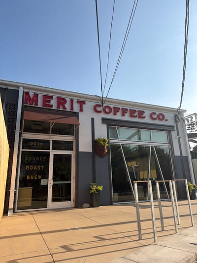 This is a photograph of the exterior of a coffee shop called "Merit Coffee Co." in Dallas, USA. The building has a modern industrial design with large windows and red raised lettering spelling out the company name. The sky is clear and blue, suggesting a sunny day. The photograph is taken from a slightly low angle, looking up at the building's facade.
On the glass doors of the entrance, text can be read, indicating the address "2639," and the words "SOURCE," "ROAST," and "BREW," along with "Merit Coffee Co. TEX. U.S.A." There are also signs for "RESTROOM" and operating hours. Several planters with greenery are visible on the exterior. Metal scaffolding or decorative elements are attached to the building above the windows. The sidewalk in front of the shop is made of concrete, and shadows from the building and surrounding structures are cast upon it. The overall impression is of a contemporary and inviting coffee shop on a bright day.