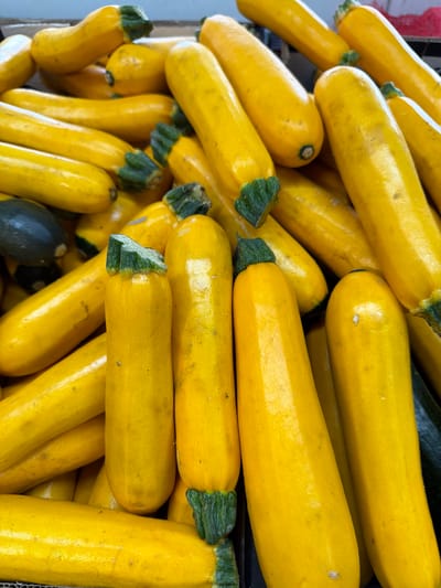 The image shows a close-up view of a pile of yellow zucchini, likely on display for sale. The zucchini are elongated and cylindrical, with a bright yellow skin. The stems are still attached and are green. A few darker green zucchini are also visible among the yellow ones. The image appears to be taken in a market or produce display setting. It is mentioned that the location is Kadima-Zoran, Israel, suggesting the photograph was taken in a market or farm in that city.
