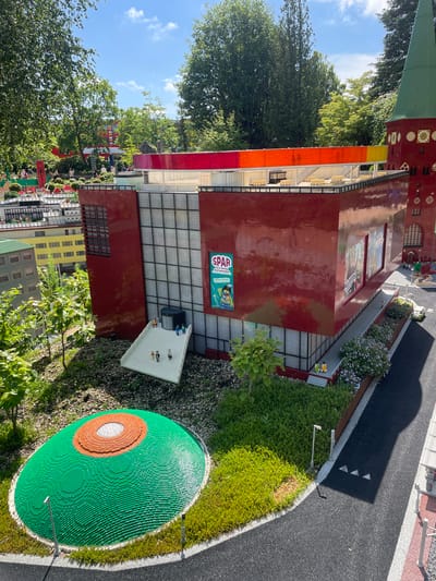 The image shows a detailed miniature model of a building and its surroundings, likely part of LEGOLAND Billund, Denmark. The central focus is a modern, multi-story building with a red brick facade and a large glass-paned section. A ramp leads up to the entrance, where several small figures, appearing to be Lego people, are positioned. Adjacent to the building is a large, circular green area with orange and white concentric circles, resembling a stylized plant or abstract design, also constructed from Lego bricks.

The surrounding environment is meticulously recreated with miniature trees, bushes, and landscaping, all made of Lego. In the background, other Lego structures are visible, including buildings and a tower with a green conical roof. A bright, multi-colored rainbow-like structure spans between two sections of buildings. A road or pathway made of dark grey Lego bricks runs along the bottom right of the image, with white lines and a few small white triangular markings. The sky above is blue with scattered white clouds, and sunlight illuminates the scene, casting shadows. There are no discernible people interacting in real-time; the figures are part of the static model.