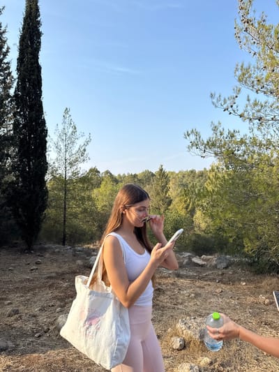 The image depicts a natural, forested outdoor environment under a clear blue sky, likely in the late afternoon or early morning due to warm lighting.

A female individual, appearing young, stands centrally. She possesses long, straight, brown hair and wears a white tank top and light pink leggings. A light-colored canvas tote bag, featuring a pink graphic of crossed tennis rackets and the text "BEVERLY HILLS ATHLETICS," is slung over her left shoulder. She holds a white smartphone vertically in her left hand and a pair of dark-lensed sunglasses in her right hand, positioning them near her face while looking at the phone screen.

In the lower right corner, the bare forearm and hand of a second individual are visible, extending into the frame. This hand holds a clear plastic water bottle with a green screw-on cap, appearing to offer it.

The ground consists of dry, uneven earth with scattered small rocks and sparse, light brown vegetation. Tall, dark green columnar evergreen trees are prominent on the left side of the frame. On the right, pine trees with visible pinecones on their branches are present. The background is composed of a dense forest of various green trees and shrubs. The sky above is uniformly light blue and clear.

This scene is consistent with a natural, forested area or green space within the vicinity of Modi'in-Maccabim-Re'ut, Israel, characterized by Mediterranean flora and terrain.