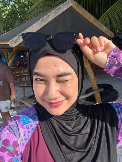 A woman wearing a black hijab and a floral swimsuit is taking a selfie outdoors. She is winking and holding her sunglasses above her head. She is standing on a beach in front of a wooden structure with a thatched roof, possibly a hut or shelter. Palm trees are visible in the background. The woman's face is illuminated by sunlight, and she has makeup on. The environment appears to be tropical.