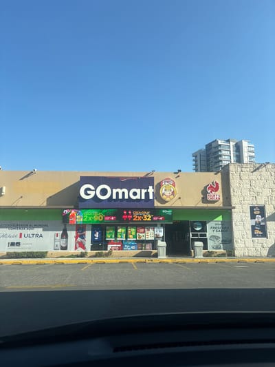 The image depicts the storefront of a "GOmart" convenience store located in Santiago Yancuitlalpan, Mexico. The store signage prominently features the GOmart logo in large, vertically oriented letters. Adjacent to the store is a "Ricky's Chicken" restaurant. Multiple advertisements are visible, including Coca-Cola promotions and Michelob Ultra beer. A multi-story building is visible in the background.