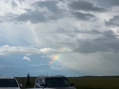 The image depicts a cloudy sky with a faint rainbow visible in the center. In the foreground, the tops of two cars are visible, with one parked to the left and another to the right. A small evergreen tree stands in the middle ground, and a fence-like structure is behind it. The background consists of a grassy field with some distant utility poles. The scene appears to be set in an open, rural area, possibly on the outskirts of Елимбетово, Russia.