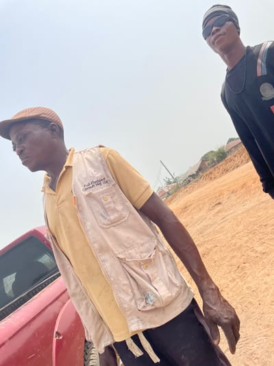 The image depicts an outdoor scene in Osara Gada, Nigeria, characterized by reddish-brown, unpaved earth. The sky is light and hazy.
In the foreground and left of the frame, an adult male with dark skin wears a brown flat cap, a yellow polo shirt, and a light beige utility vest. The vest displays "Full-Fledged Consult Nig. Ltd." on the left chest area, has multiple pockets, and an open zipper. He looks towards his left (viewer's left), with a visible, aged left hand and forearm. A portion of a red vehicle, likely a truck or SUV, is partially visible behind him on the far left.
In the background and right of the frame, a second adult male with dark skin is visible. He wears a black beanie, black rectangular sunglasses, and a long-sleeved black shirt with design elements on the shoulder. A black beaded necklace is around his neck. He looks forward and slightly to his right (viewer's right).
The background beyond the individuals includes distant structures, possibly houses with corrugated roofs, sparse vegetation, and a utility pole, suggesting a rural or semi-urban setting. No direct interaction between the two individuals is observed; both are standing.