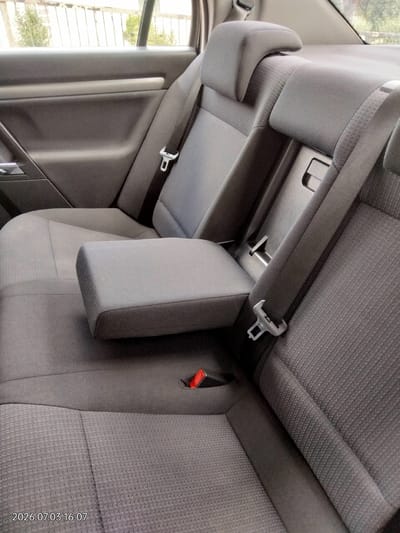 The image displays the interior of a car's back seat, featuring grey fabric upholstery. The seat includes two headrests, a folded-down armrest in the center, and seatbelts. The armrest is positioned over the seat. The background shows the interior door panel of the car, with a window revealing a blurry outdoor scene. The car's interior is clean and appears to be in good condition. There are no people or animals present in the image. The timestamp in the lower-left corner indicates the photo was taken on 2026.07.03 at 16:07. This scene is located in Novyi Myliatyn, Ukraine.