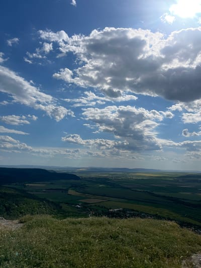 The image depicts a landscape scene. In the foreground is a field of green grass, suggesting a hillside or edge of a plateau. Beyond the foreground, a valley with agricultural land is visible, including fields and scattered vegetation. In the distance, there's a hazy view of a landscape, possibly indicating distant mountains or terrain obscured by atmospheric conditions. The sky occupies a large portion of the image, filled with white clouds and a bright sun. The overall impression is one of a high vantage point overlooking a rural landscape on a sunny day.
There is insufficient information in the image to determine the specific location as Razdelna, Bulgaria.