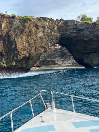 A forensic examination of the image reveals a maritime scene near the city of Sakti, Indonesia. The foreground displays the white bow of a boat, equipped with polished metal railings on either side and a central silver-colored cleat. Light blue, textured non-slip pads are visible on the boat's deck. The boat is positioned on a body of deep blue-green water, exhibiting ripples and small waves.

In the midground, to the left, white-capped waves are observed breaking against the base of a substantial rock formation. The background is dominated by a large, dark brown and gray rocky cliff, characterized by a prominent natural archway in its central section. Through this archway, a lighter-colored sandy beach area is discernible, with calmer water approaching it. The rock face displays varied textures and erosion marks, with some lighter white deposits, particularly within the arch. Sparse green vegetation is visible along the top edge of the cliff against a pale blue sky with scattered clouds. No individuals are visible in the frame.