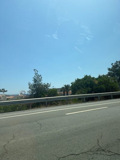 This media file depicts a roadside scene in Parekklisia Community, Cyprus, under a clear, bright sky, suggesting a sunny day. The primary focus is a cracked asphalt road with a white line marking the lane. To the left of the road, a metal guardrail separates it from a lush green area characterized by trees and vegetation.
In the background, beyond the greenery, buildings are visible, hinting at a semi-urban or coastal settlement. Among the trees, a palm tree stands out. The sky is a vibrant blue, with some reflections visible on what appears to be a window or a reflective surface in the upper left portion of the frame, possibly from the perspective of a vehicle. The image is oriented sideways, with the road running diagonally from the bottom right to the top left of the frame.
No people or specific activities are clearly discernible, but the scene evokes a sense of travel or a journey, given the road and the perspective. The weather appears to be pleasant and warm, typical of Cyprus. The cracked asphalt and guardrail suggest a well-traveled road, likely a public highway. The combination of road, greenery, and distant buildings paints a picture of a typical Mediterranean landscape. No text is visible in the image.