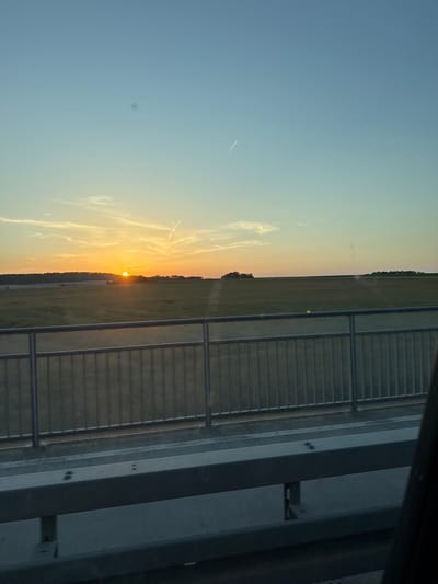 The image shows a sunset over a rural landscape, viewed from what appears to be a bridge or overpass. The sky is a gradient of pale blue to warm orange, with the sun just above the horizon. Wispy clouds are visible, catching the golden light of the setting sun. Below the sky is a vast expanse of green fields, with a line of trees forming a dark silhouette in the distance. A metal railing runs horizontally across the foreground, suggesting the vantage point is elevated. The overall impression is peaceful and expansive. This scene is not specific to Giessen, Germany, as it depicts a generic rural setting that could be found in many locations.