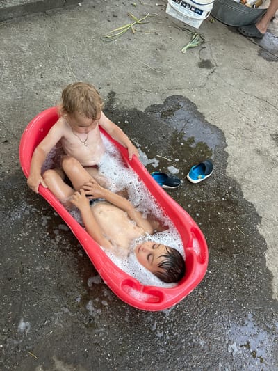 Two young boys are bathing in a red plastic tub filled with soapy water on a concrete surface. The tub is positioned outdoors, with scattered debris and puddles of water on the ground. A pair of blue and black water shoes are placed next to the tub. The scene appears to be a casual, perhaps backyard, setting in Chernove, Ukraine.