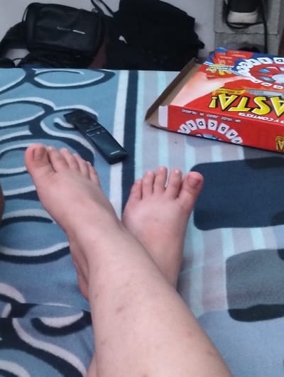 A photo shows crossed legs and feet on a patterned blanket, with a remote control nearby. A red and white game box sits on the blanket as well. In the background, a black bag or backpack is visible, along with a light-colored shelf or surface. The setting appears to be a casual, indoor environment.