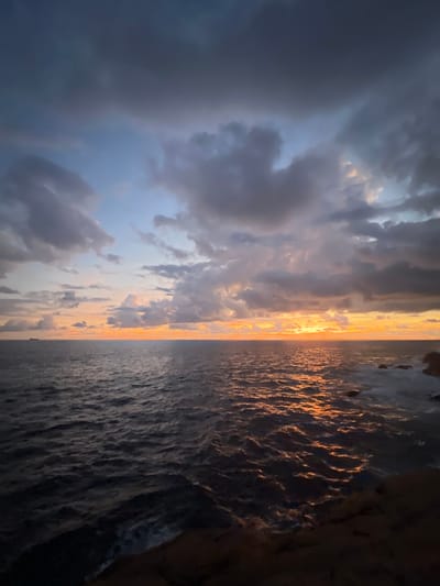 The image captures a dramatic coastal landscape scene, likely at either sunrise or sunset, from Rumassala, Sri Lanka. The photograph appears to have been taken in portrait orientation and shows a view across a vast ocean, framed by a prominent cliff on the right side.

The upper portion of the frame (when rotated correctly to landscape orientation) is dominated by a sky filled with dynamic clouds. Large, dark grey and bluish clouds cover much of the left side, indicating possible dense cloud cover. However, a brilliant golden light emanating from near the horizon illuminates the clouds in the center and right, creating warm orange and yellow hues that contrast with the cooler tones of the darker clouds. Patches of lighter blue sky are visible between the cloud formations.

A distinct horizon line separates the sky from the expansive ocean below. The water surface, particularly closer to the bright light source, reflects the golden glow, creating a shimmering, textured effect with visible ripples and waves. Further away from the direct light, the ocean appears darker, a deep grey-blue. In the far distance, a small, dark silhouette of what appears to be a ship or boat can be observed near the horizon.

On the far right of the image, a dark, rugged cliff face or rocky outcrop partially obscures the scene, suggesting the vantage point is from within or very close to a cave or an overhang. Its textured surface is seen in silhouette, adding depth and framing the vibrant sky and water. The overall impression is one of majestic natural beauty and a serene, contemplative atmosphere as daylight transitions over the Indian Ocean. No visible text is present in the media file.