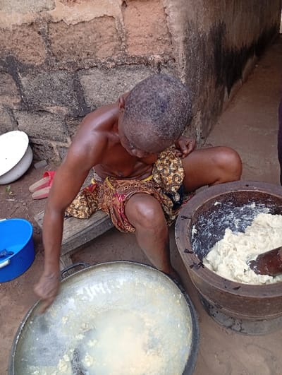 An adult individual, likely a woman, is captured in an overhead shot, engaged in food preparation outdoors in Obwe, Nigeria. She is seated on a low wooden stool, clad in a vibrant patterned wrapper in shades of red, gold, and dark brown, with her upper torso bare and skin appearing somewhat shiny, possibly from exertion or oil. Her dark hair is closely cropped.
The central activity involves her stirring a thick, light-colored, pasty substance within a large, round aluminum pot with her left hand. To her right, a substantial wooden mortar holds a similar light-colored mixture, with a heavy wooden pestle resting inside. This setup suggests the traditional preparation of staple foods like fufu or pounded yam, involving manual pounding and mixing.
The setting is outdoors, against a rustic wall constructed from rough-hewn, reddish-brown and dark grey stone or mud bricks. The ground is bare earth. Nearby, several household items are visible: a white enamel bowl and a blue plastic bucket are on the ground to her left, along with a pair of red flip-flops. A dark, narrow passageway or corner of a building is discernible in the background to the right. The scene conveys a sense of diligent work in a traditional, possibly rural, domestic environment during daylight hours. No visible text is present in the image.