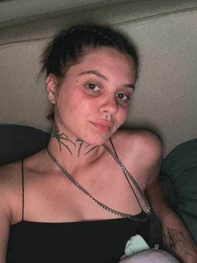 A young woman with dark hair pulled back into a bun and a nose ring is looking at the camera. She is wearing a black tank top and a silver chain necklace. She has a spider tattoo on her neck and a tree tattoo on her arm. The background appears to be a couch or bed. The lighting is dim. No location in Genemuiden, Netherlands is identifiable.