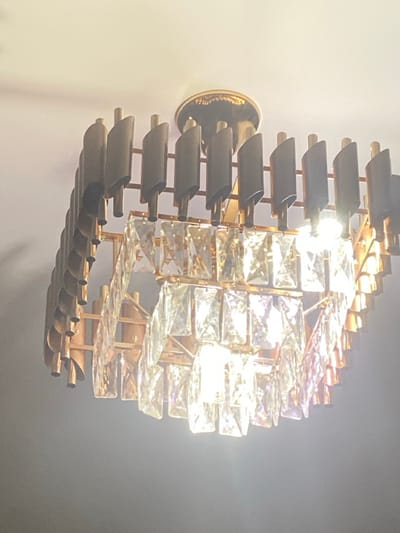 A wall-mounted light fixture, predominantly gold and crystal. The fixture features a circular design, with rows of dark, curved metal pieces emanating from a central gold base attached to the wall. Alternating rows consist of horizontal, rectangular crystals that create a dazzling, reflective effect due to the internal light source. The overall design projects a sense of modern elegance.
