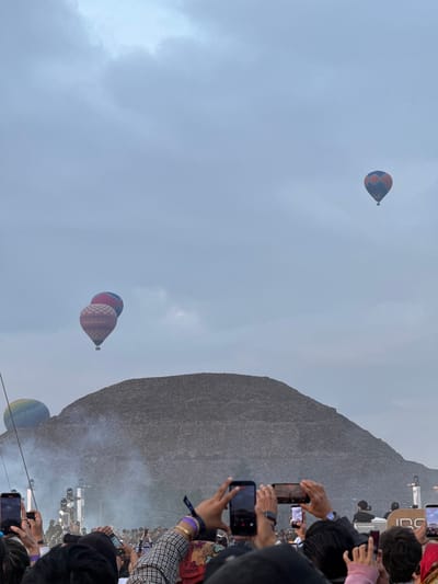 The image shows a scene in San Juan Teotihuacán, San Francisco Mazapa, Mexico. In the background, a large pyramid-like structure looms. Three hot air balloons float in the sky: one higher up with a blue and red pattern, a pair closer to the pyramid with red and blue designs, and another lower balloon with a rainbow hue. In the foreground, a crowd of people holds up phones, presumably taking pictures of the pyramid and balloons. The sky is overcast.