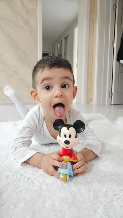 A young child is lying on a white, fluffy rug, indoors. The child, possibly male, has short, dark hair and is wearing a white long-sleeved shirt. The child's mouth is open, tongue extended, and eyes wide. The child is holding a Mickey Mouse toy in their hands. The background includes a tiled wall and a partially visible doorway leading to another room. The location appears to be a private residence. The scene is unlikely to be in Iasenovets, Bulgaria.
