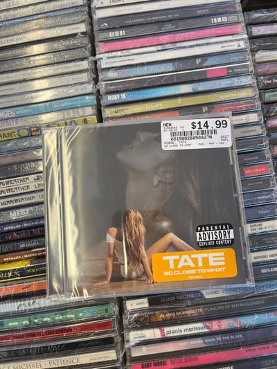 The image displays a retail environment featuring a dense array of compact discs (CDs) on display, located in Edmond, United States. The central focus is a new, shrink-wrapped audio CD titled "TATE SO CLOSE TO WHAT" by MCRAE, priced at $14.99, as indicated by a white label with a barcode, artist, and album title (MCRAE, TATE SO CLOSE TO WHAT MUS - POP - MUCL) and a visible date of 02/24/25. The album cover features two overlapping images of a woman: a faint, ghosted profile looking upward in the background, and a more prominent, seated figure with long, light-colored hair, wearing an off-shoulder garment, looking away from the viewer. A black and white "PARENTAL ADVISORY EXPLICIT CONTENT" label is visible on the lower right of the album art. This central CD rests horizontally atop a stack of other shrink-wrapped compact discs. The surrounding area is densely populated with similar stacks of CDs, arranged in horizontal rows, displaying numerous album spines from various artists including Kylie Minogue, Moby, Metallica, Alanis Morissette, and Gary Moore. The scene is illuminated by interior lighting, casting reflections on the plastic CD wrappers. No individuals are physically present within the frame; only their images are depicted on the album covers.