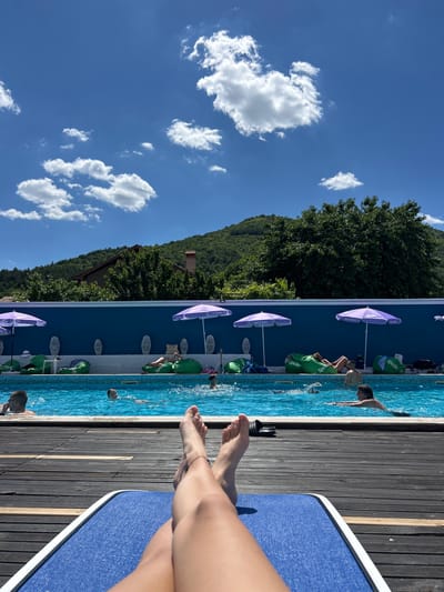 The image depicts a sunny day at an outdoor swimming pool, likely located in Varvara, Bulgaria, given the context. In the foreground, the legs and feet of a person are visible, resting on a blue lounge chair situated on a wooden deck. Beyond the deck, the clear blue water of the swimming pool is the central focus. Several people are in the pool, with some swimming and others relaxing.
Along the edge of the pool, a vibrant blue wall serves as a backdrop. Several large green beanbag chairs and purple umbrellas are arranged along this wall, suggesting a recreational area. In the distance, lush green trees and a forested hillside rise up, with a house visible nestled amongst the trees. The sky above is a brilliant blue, dotted with fluffy white clouds, indicating fair weather. The overall scene conveys a sense of relaxation and enjoyment on a summer day.